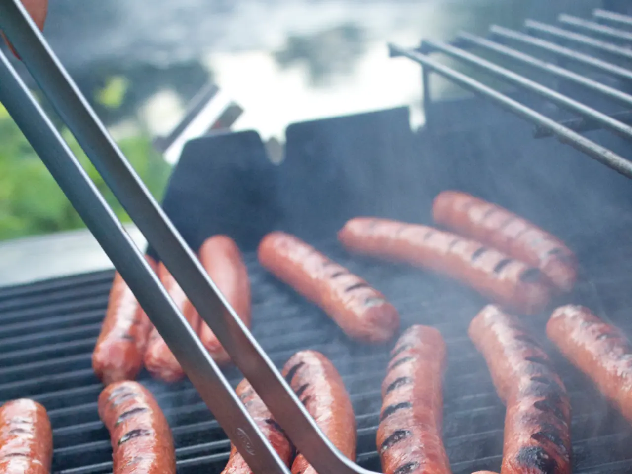 In this image I can see sausages on a grill and tongs. In the background I can see a person's hand...