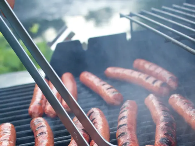 In this image I can see sausages on a grill and tongs. In the background I can see a person's hand...