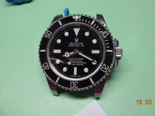 In this picture we can see a watch dial and a paper on a green platform and at the bottom right...