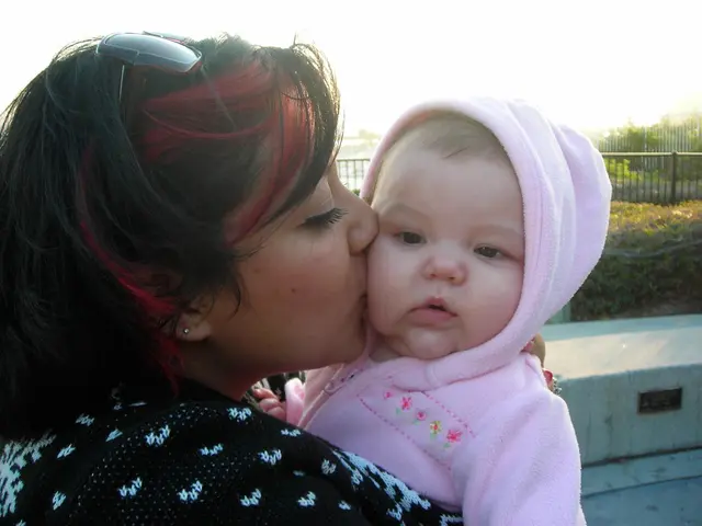 In the center of the image a lady is hugging a baby. In the background of the image we can some...